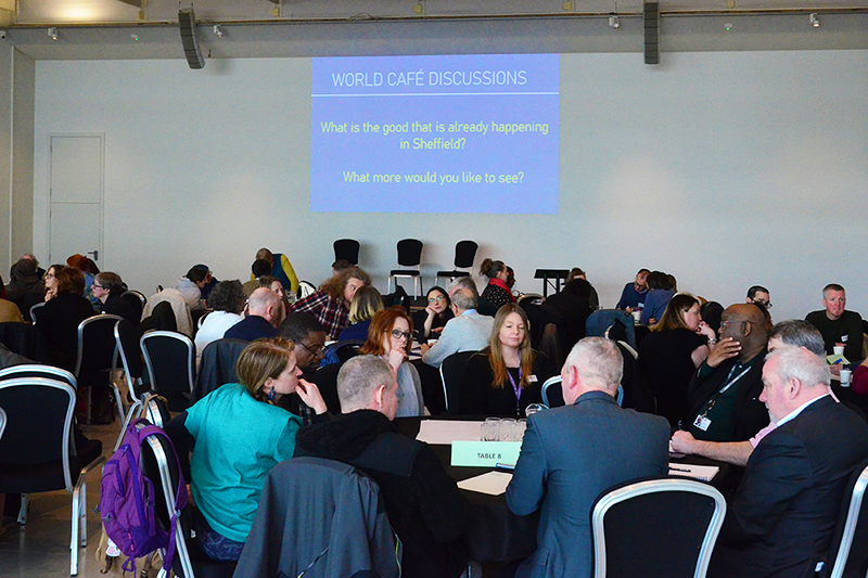 People sat round tables at an event. The big screen says "world cafe discussions. What is the good that is already happening in Sheffield. What more would you like to see?"